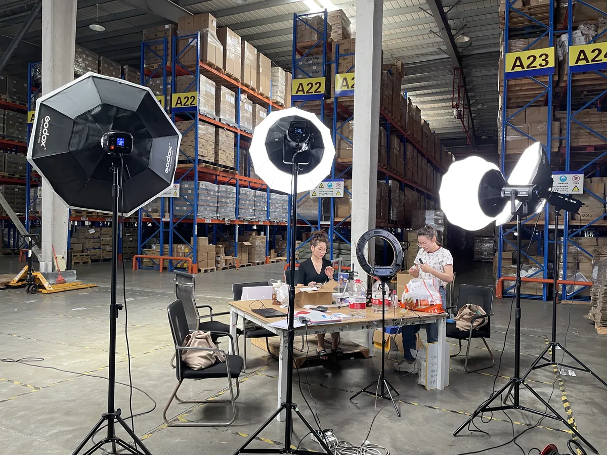 On-site live broadcast inside the China bonded zone: octagonal key lights, presenters at a desk, A22 / A23 high-bay racks in the background — proving inventory is real and ships from China.